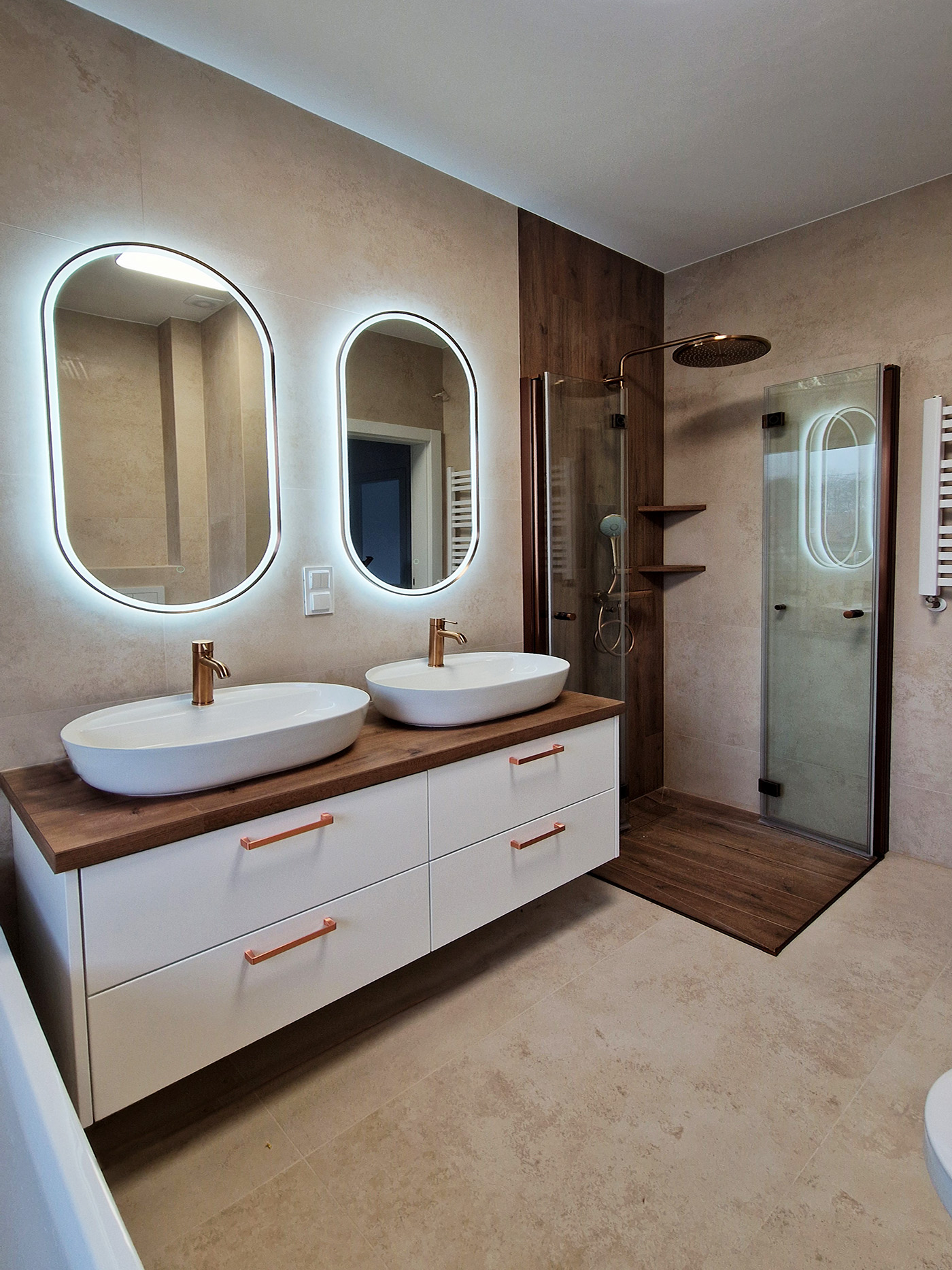 Nowoczesna łazienka w jasnych beżowych płytkach z drewnopodobnym wykończeniem pod prysznicem, dwiema umywalkami, wanną oraz owalnymi lustrami z podświetleniem LED i armaturą w kolorze szczotkowanej miedzi - Łomianki